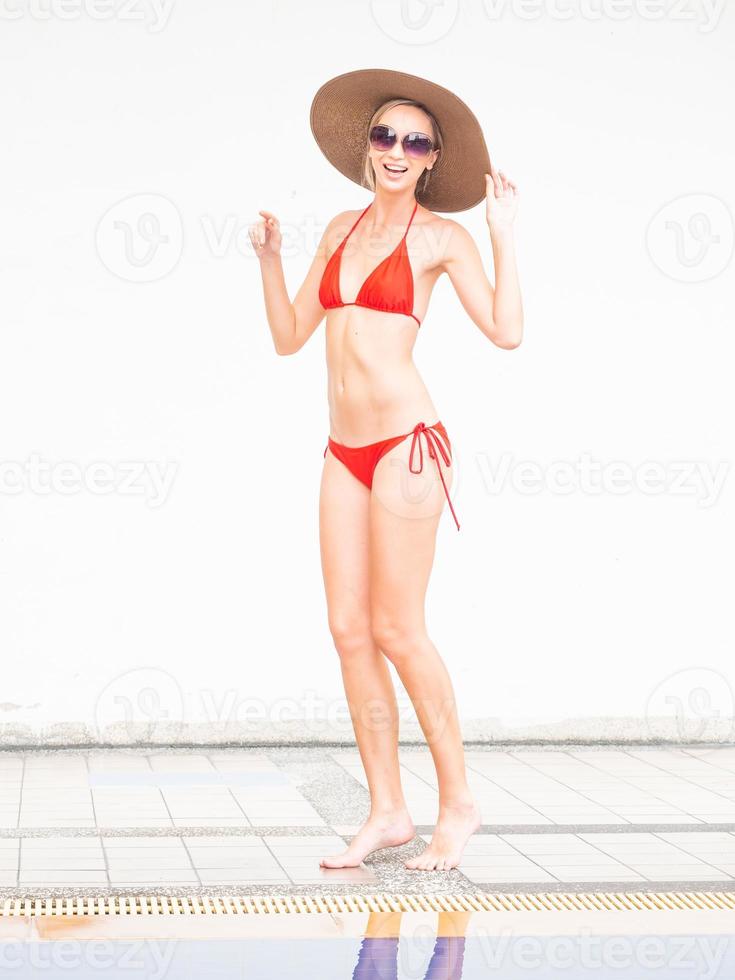 chica sexy chica feliz en bikini rojo usa sombrero y gafas de sol junto a la piscina con pared blanca. foto