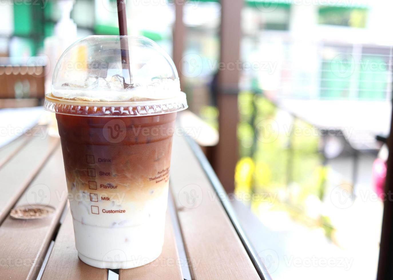 café helado dos capas de café y leche en un vaso de plástico cerrado con paja sobre una mesa de color marrón oscuro y desenfoque de fondo brillante en tailandia. foto