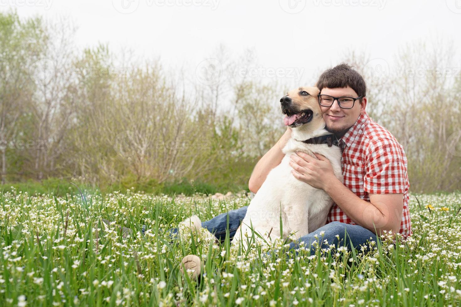 Happy man sitting with mixed breed shepherd dog on green grass in spring flowers photo