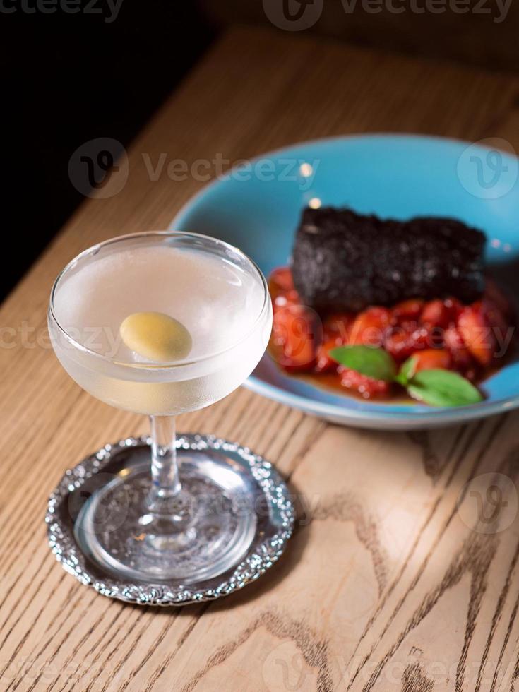 lomo de cerdo al horno marinado en tinta de sepia con tomates rojos y albahaca en plato azul, sal, pimienta, cubiertos y cóctel de alcohol de vermú seco con oliva en la mesa del bar foto