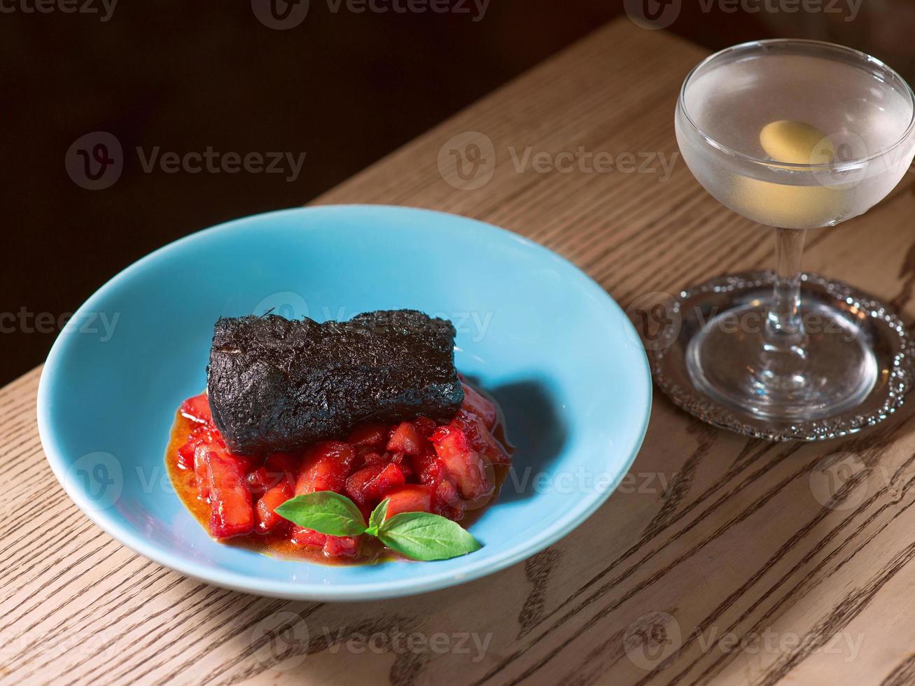 lomo de cerdo al horno marinado en tinta de sepia con tomates rojos y albahaca en plato azul, sal, pimienta, cubiertos y cóctel de alcohol de vermú seco con oliva en la mesa del bar foto
