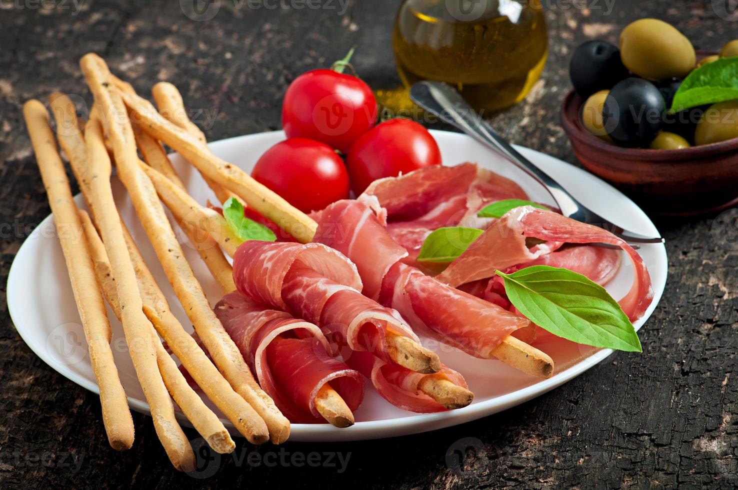 Grissini bread sticks with ham, olives, basil on old wooden background photo