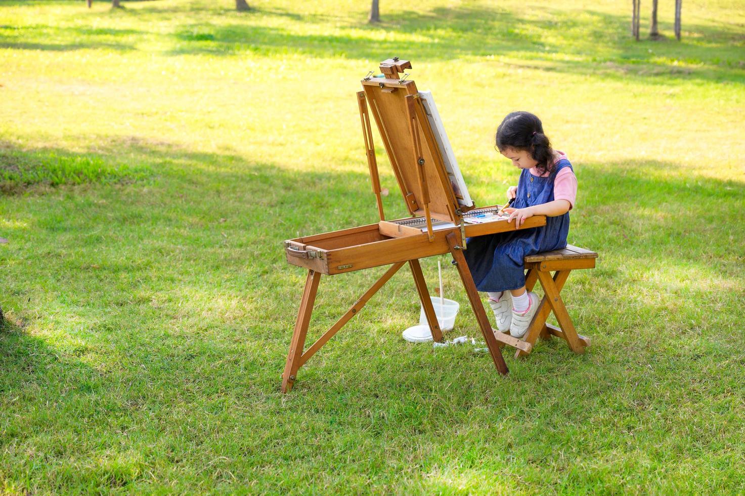 A little girl is sitting on the wooden bench and painted on the canvas placed on a drawing stand photo