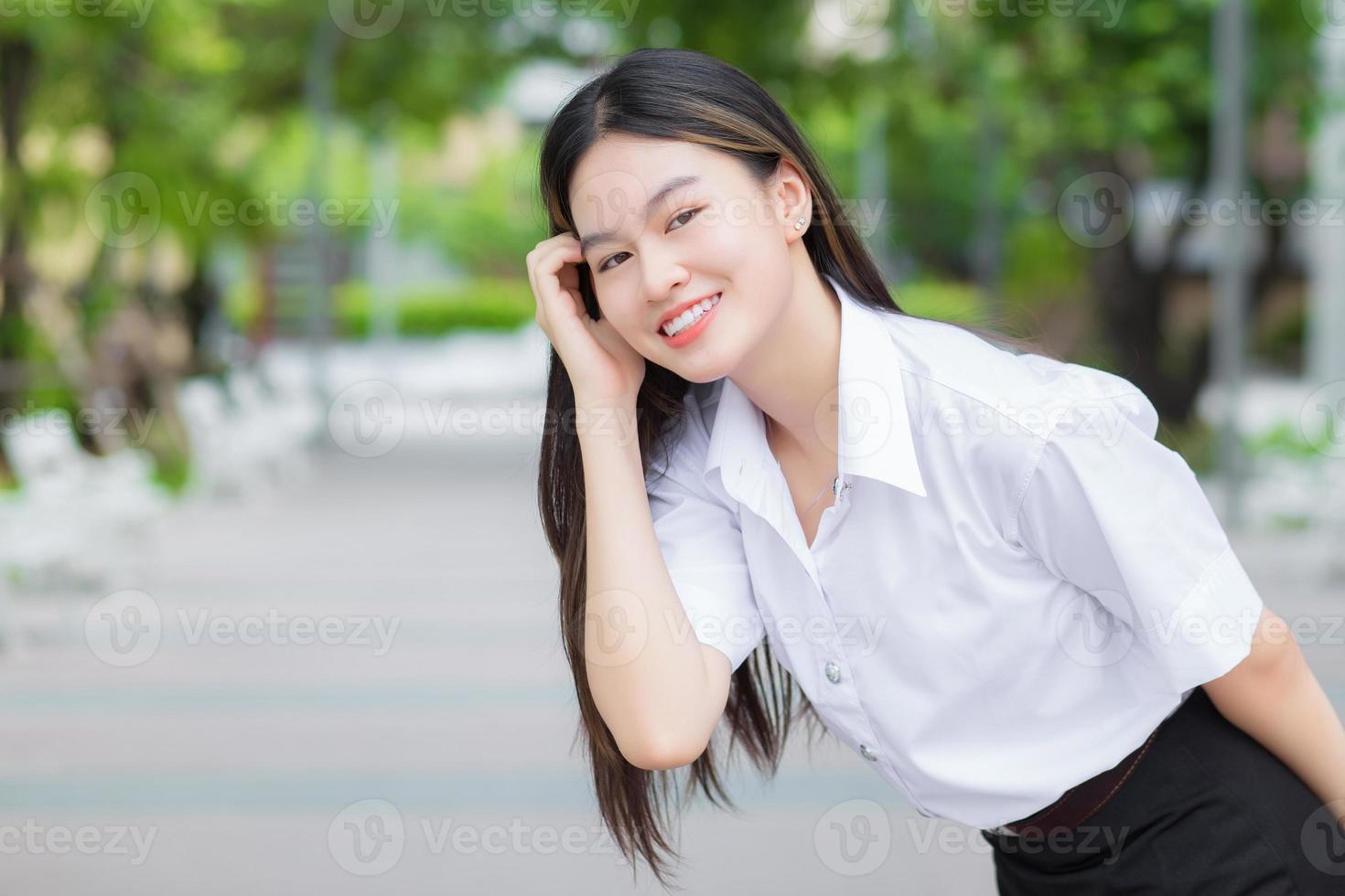 Young Asian woman student who has blond long hair smiles and looks at to camera while holding hands and standing in university with outdoor garden tree background. photo