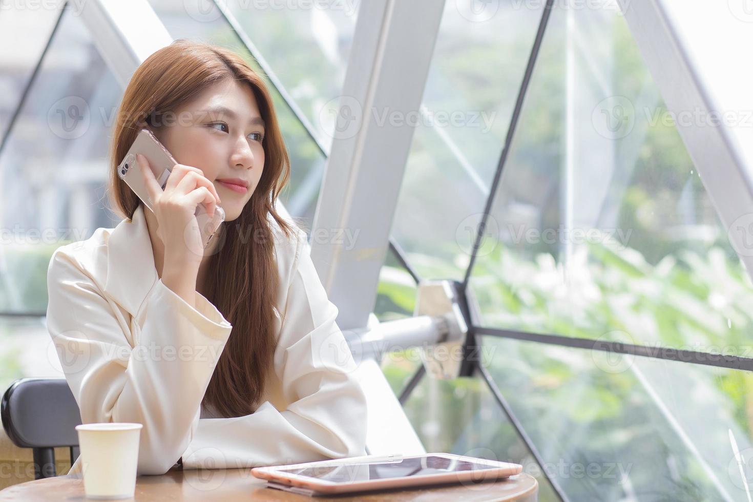 Beautiful young Asian professional business female in a white shirt is  while using your smartphone in your hand to talk she is sitting working at coffee shop among business. photo