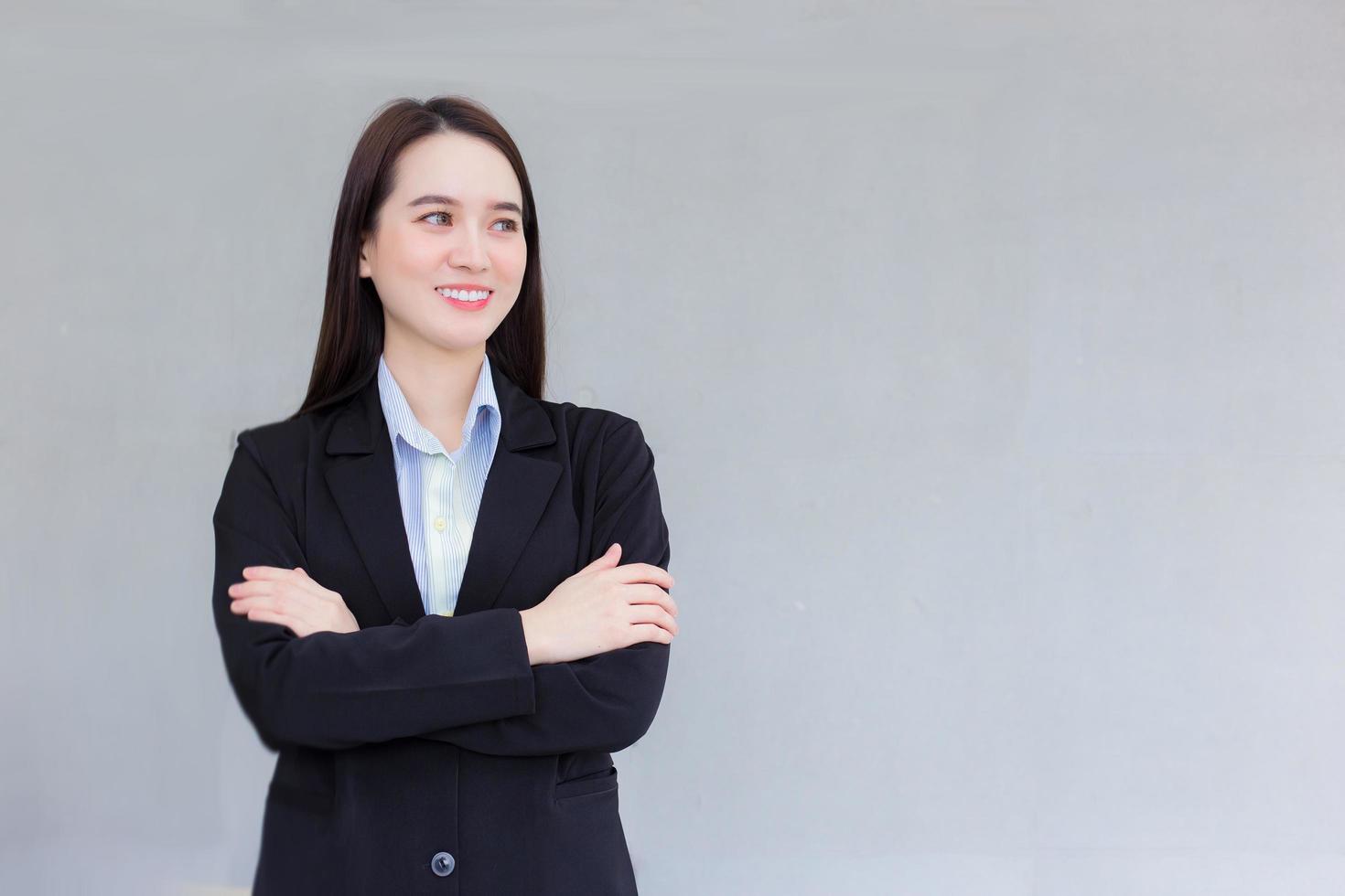 Professional business working Asian woman who wear a black formal suit stands with crossing her arm smiles happily at office as background. photo