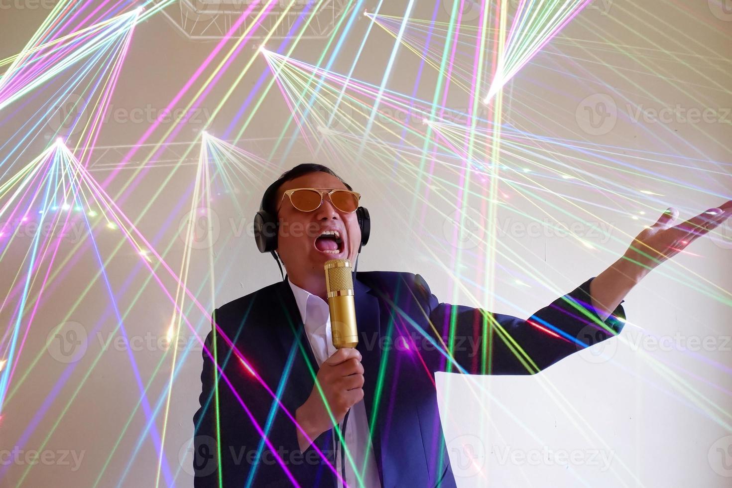 Portrait of the handsome attractive singer in a suit and brown glasses, singing hit with open mouth in gold microphone gesture with hand isolated on grey background and shine color light. photo