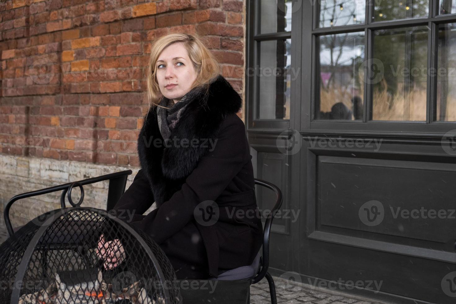 Blonde in coat with fur collar sits at table and warms her hands over glowing coals. photo