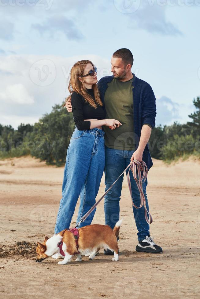 una joven pareja feliz y un perro se paran en la playa contra pinos y arena. hombre guapo y mujer hermosa foto