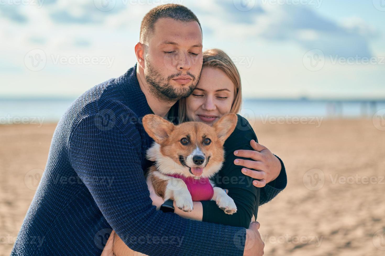 Young happy couple with dog standing on beach. Beautiful girl and guy and Corgi puppy having fun photo