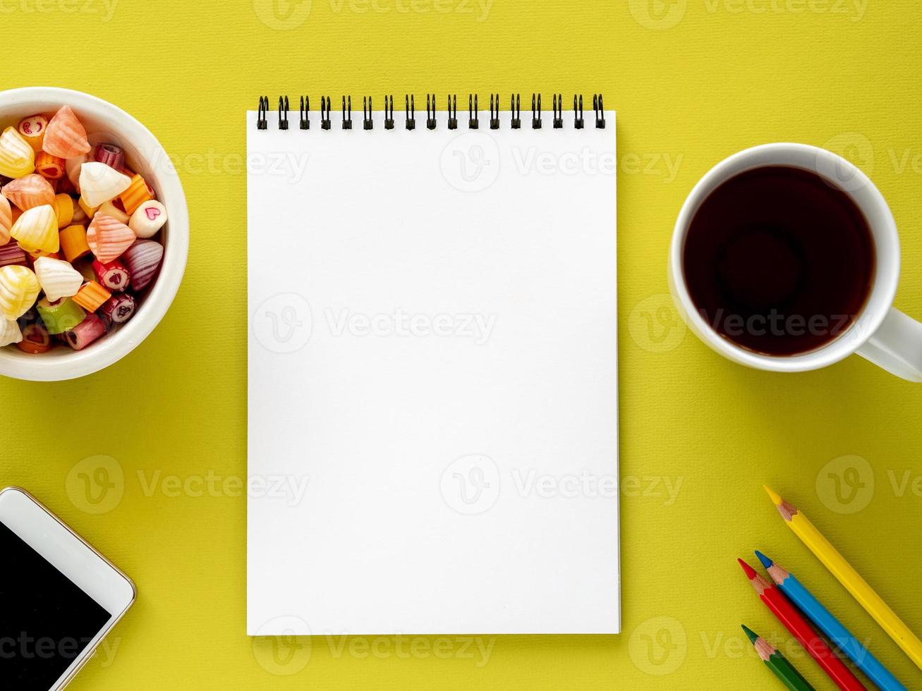 Open notepad on spiral with clean white page, cup with tea, caramels in a bowl, smartphone and color pencil on bright yellow background color photo