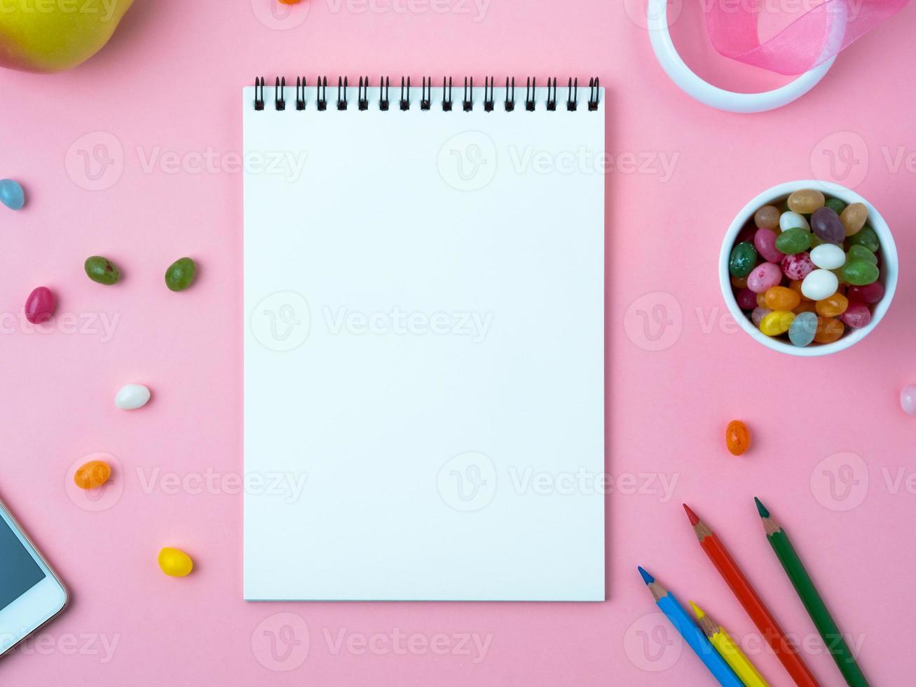 Open notebook with a clean white sheet, sweets, mobile phone, crayon, decorations on a pink bright table photo