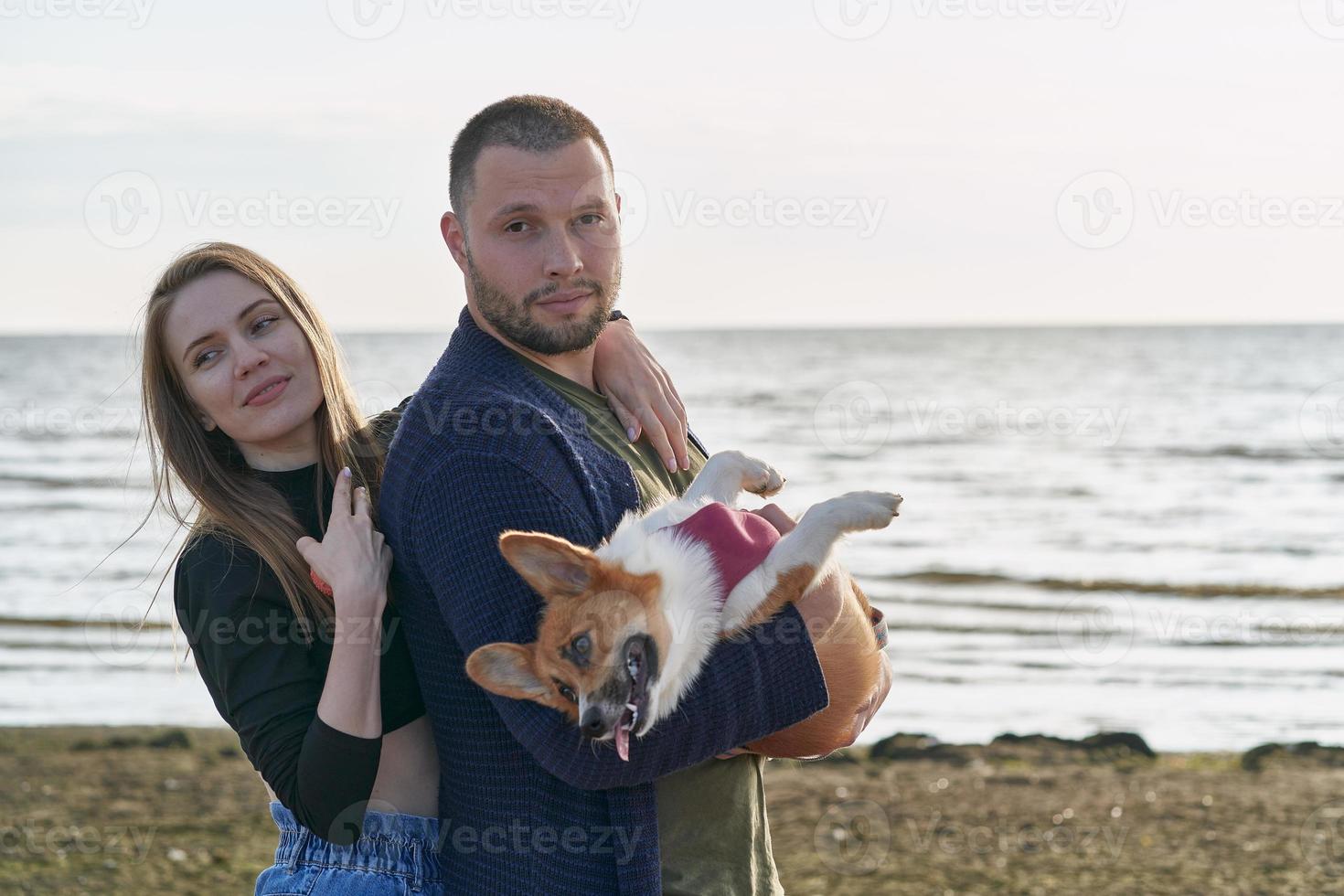 Young happy couple with dog standing on beach. Beautiful girl and guy and Corgi puppy having fun photo