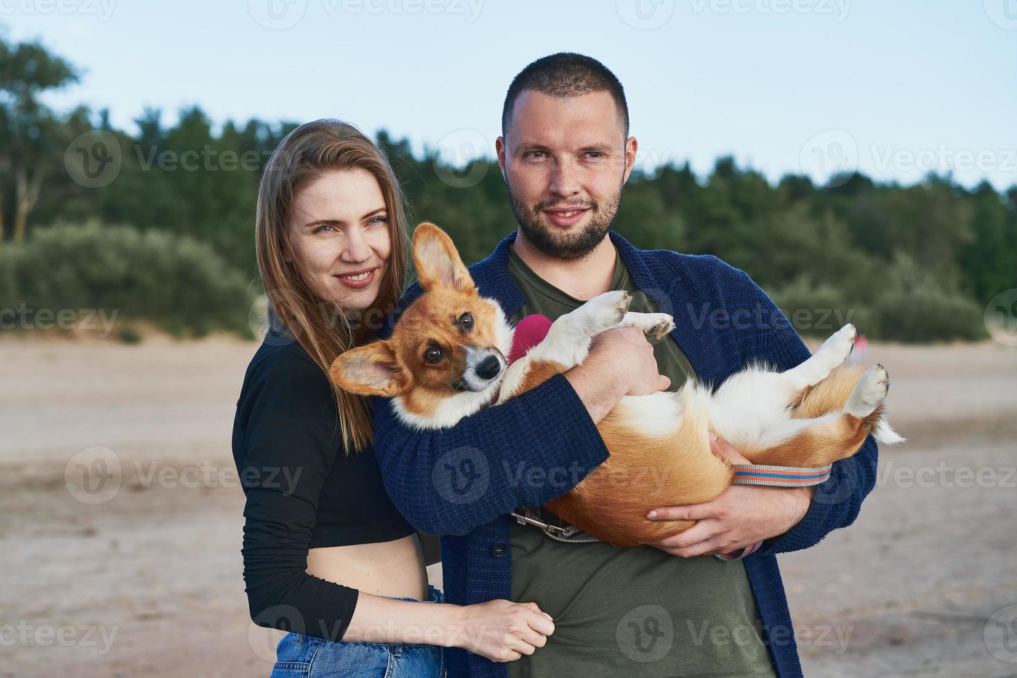 Young happy couple with dog standing on beach. Beautiful girl and guy and Corgi puppy having fun photo