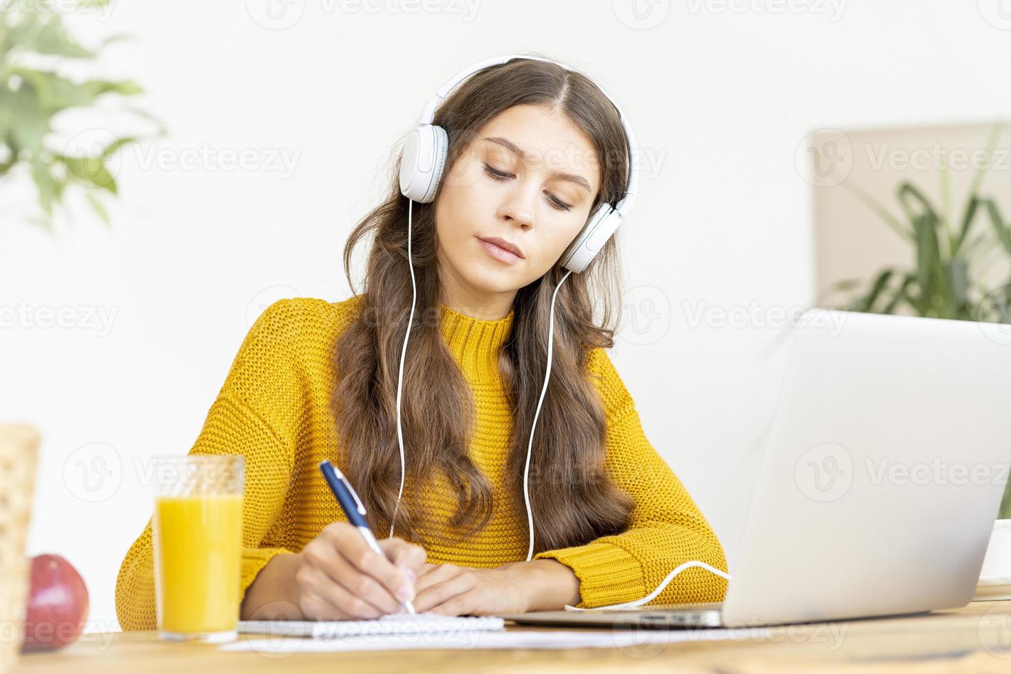 Happy female in wireless headphones studying online course, using pc and writing in notepad photo