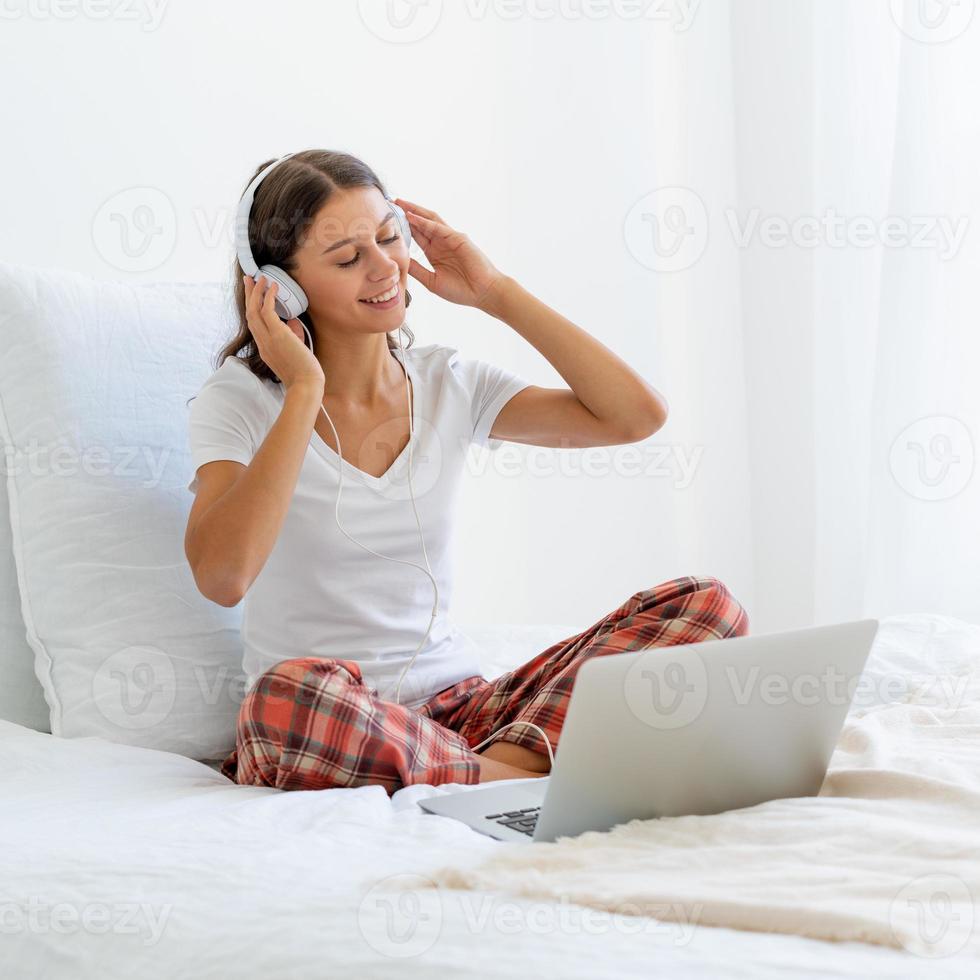 Young smiling woman sitting on bed in bedroom and listening to music or watching movie photo