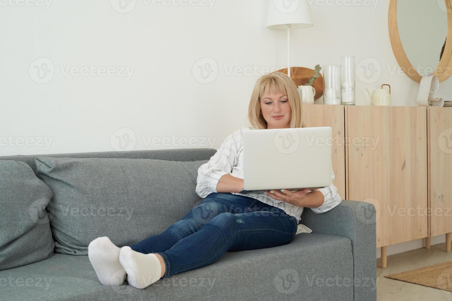 Mature woman using laptop while sitting on comfortable sofa, home interior photo