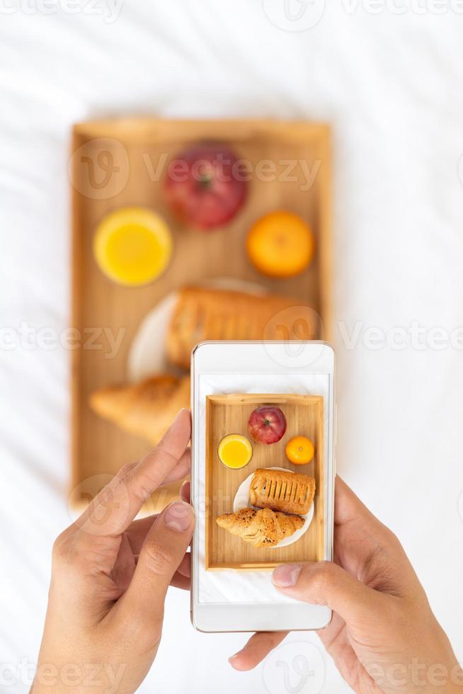 Blogger taking photos of food, shooting Breakfast in bed at hotel on mobile phone, tray with juice