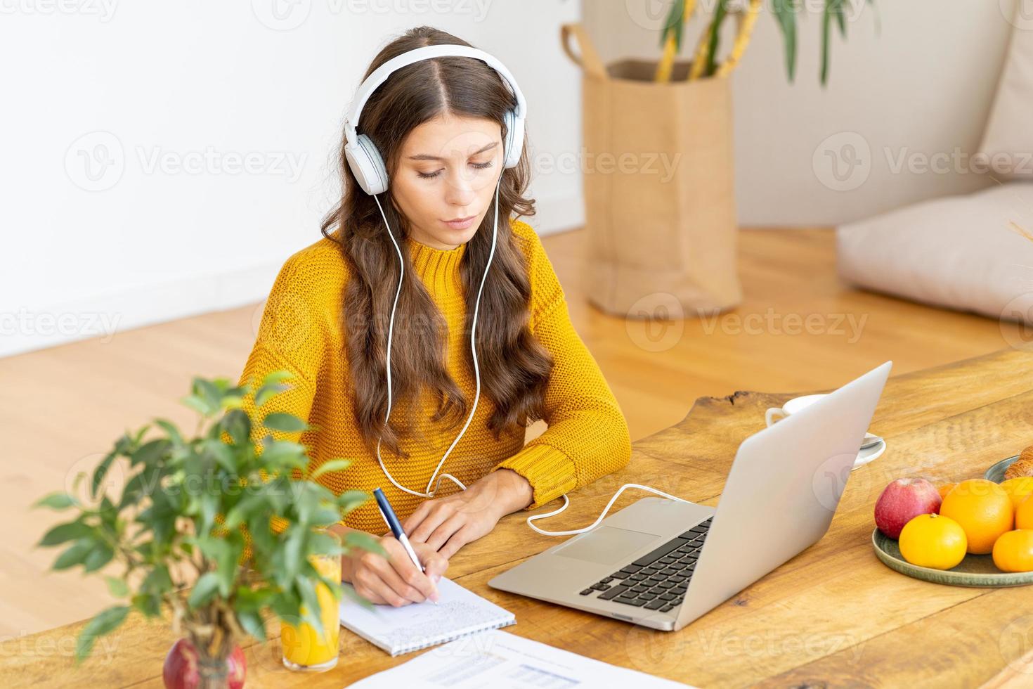 Happy female in wireless headphones studying online course, using pc and writing in notepad photo