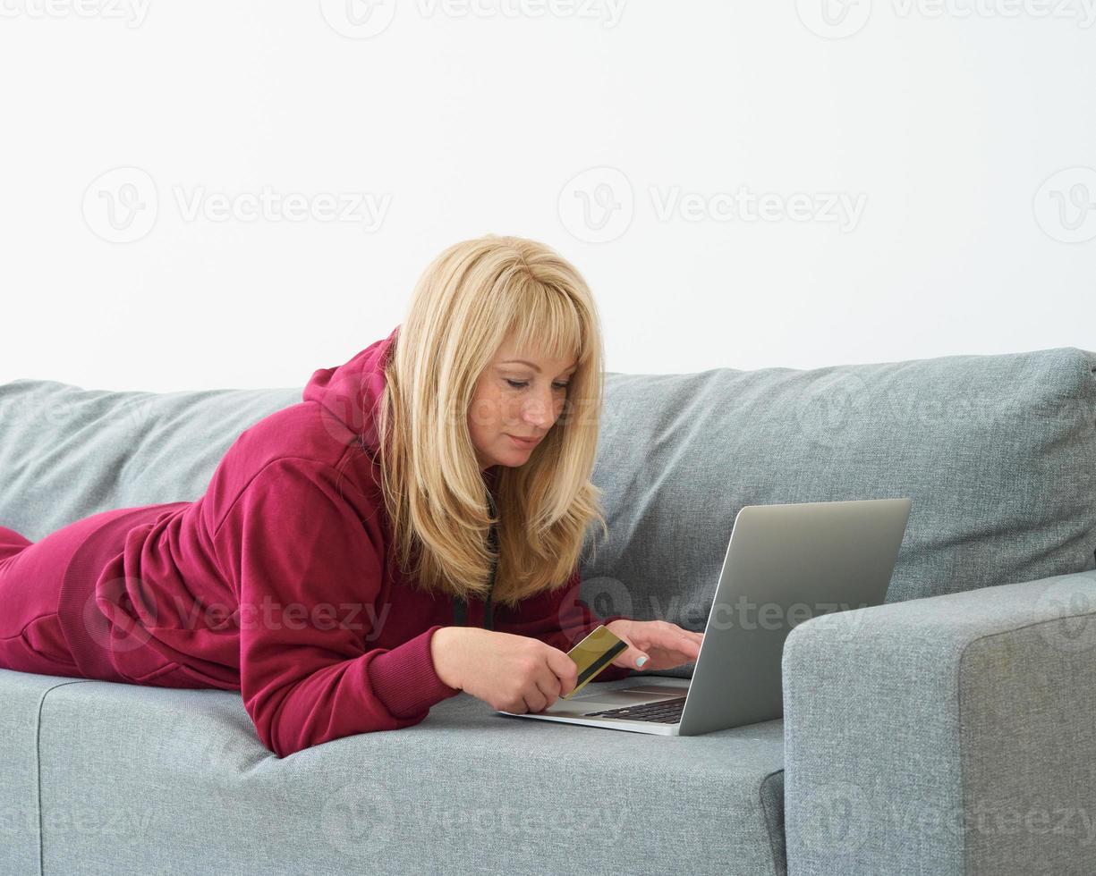 Mature woman online shopping, lying on couch with laptop and credit card. Middle-aged female photo