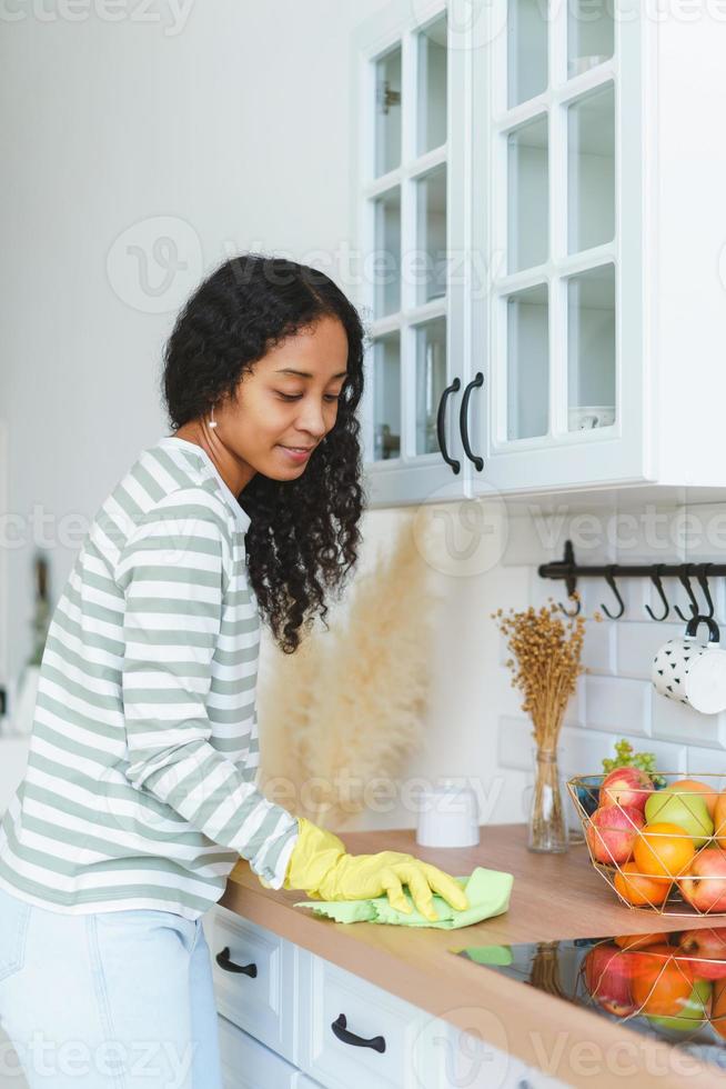 vertical de una mujer afroamericana sonriente lavando la superficie de la cocina con guantes y una toallita foto