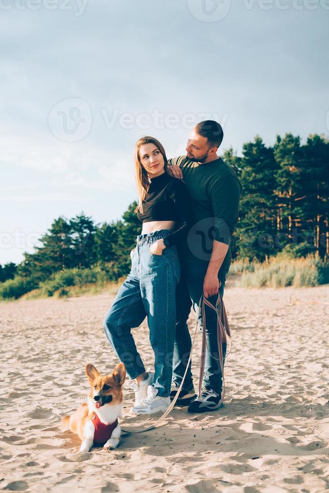 Young happy couple and dog standing on beach against pines and sand photo