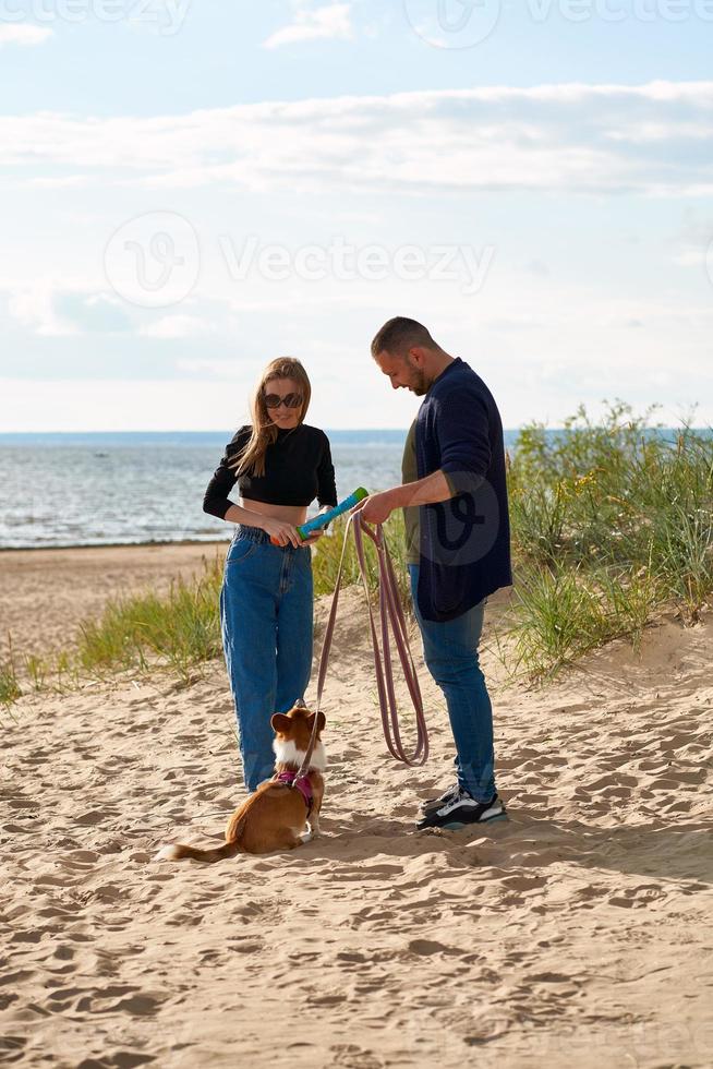 Young happy couple playing with dog by toy. People walking along beach with pet. photo