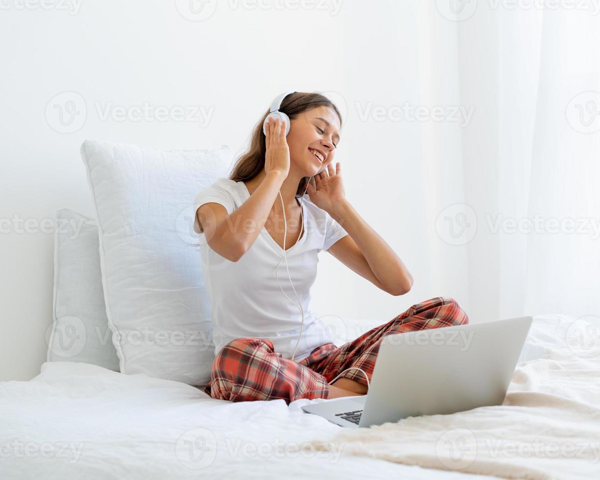 Mujer sonriente joven sentada en la cama en el dormitorio y escuchando música o viendo una película foto