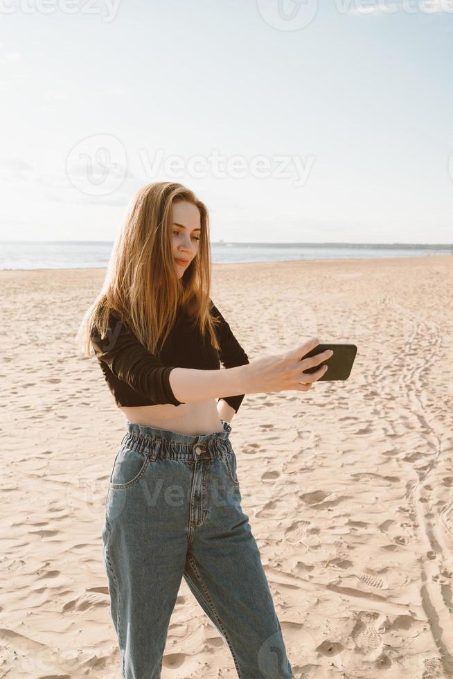 Pretty female with long hair, blonde takes photo on mobile phone on sandy beach in summer