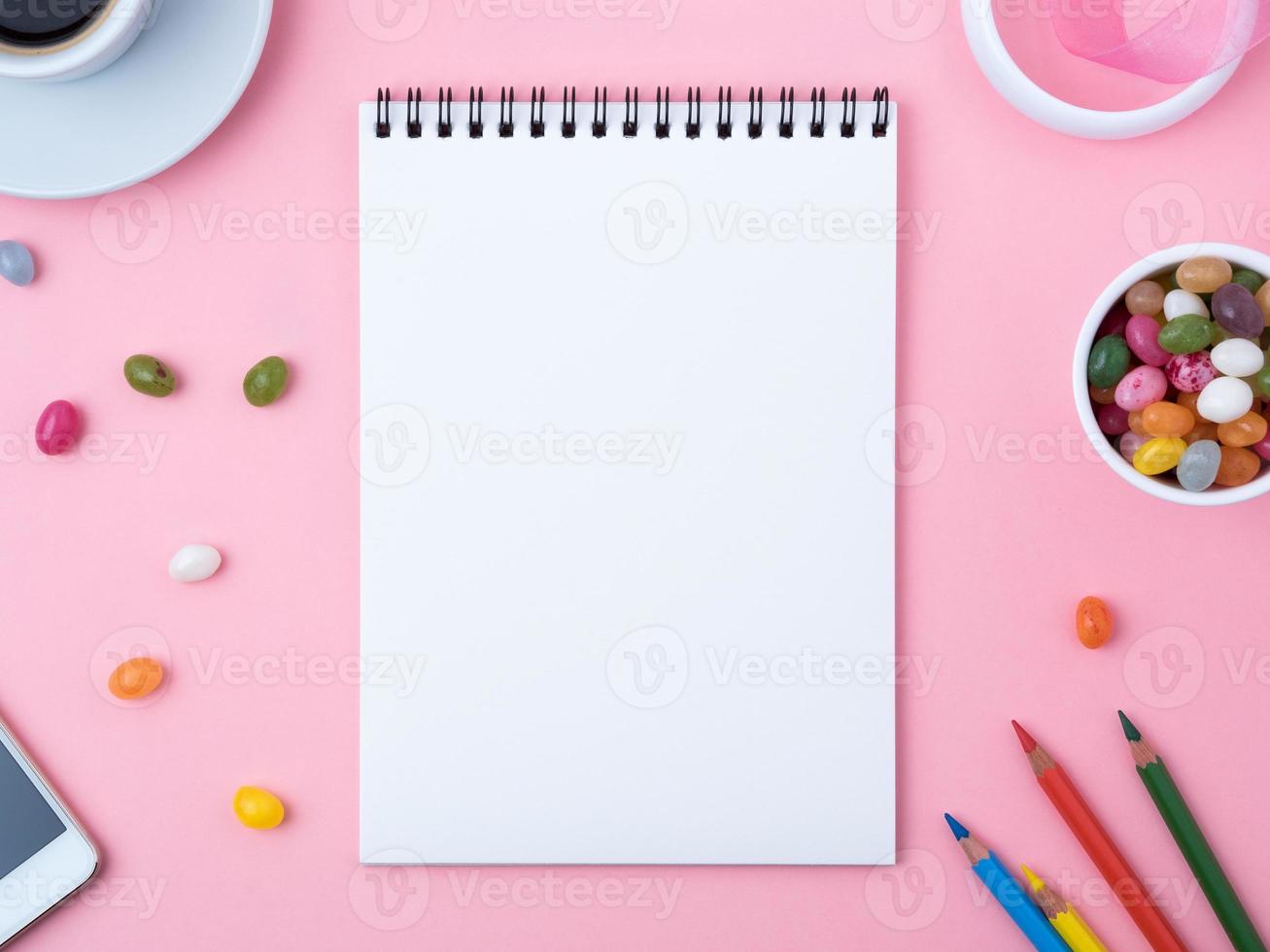 cuaderno abierto con una sábana blanca limpia, caramelo, piruletas, teléfono móvil, crayón, taza de café, decoraciones en una mesa rosa brillante. el lugar de trabajo de las niñas para la creatividad, los planes y los sueños. foto