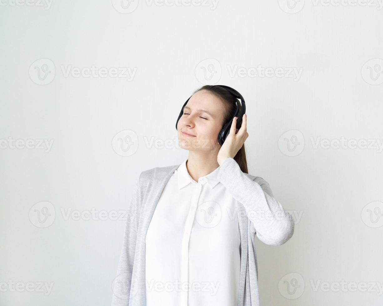 Young female is listening to music with headphones, girl standing in front of light gray wall photo