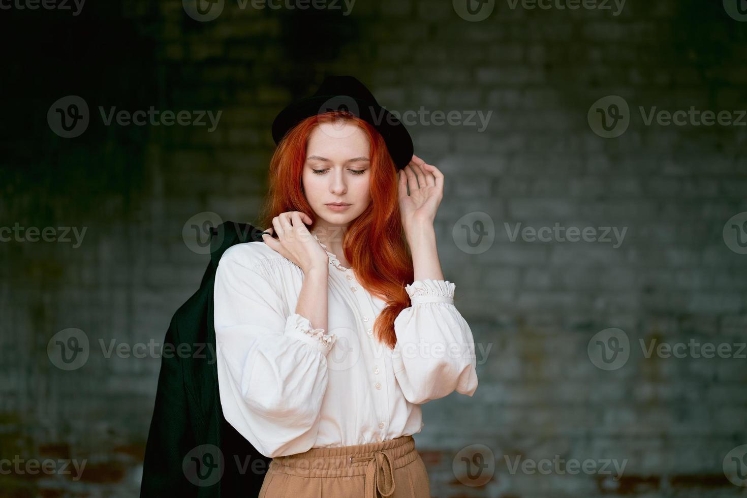 Redhead woman is resting against dark grunge wall. Female in black hat photo