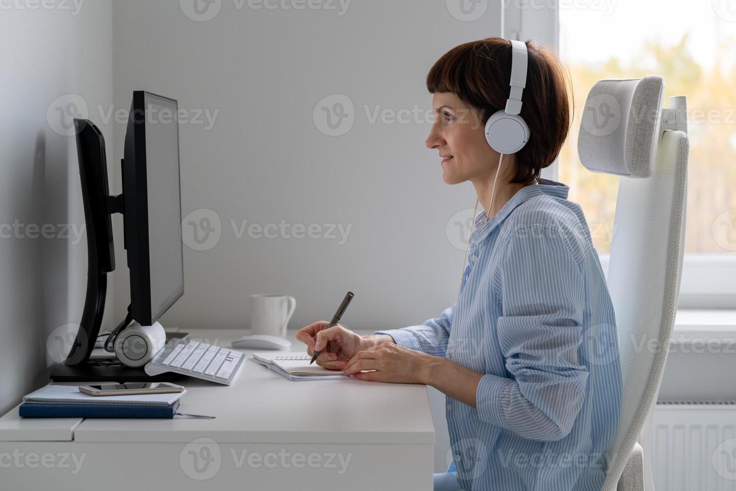 Happy female in wireless headphones studying online course, using pc and writing in notepad, photo