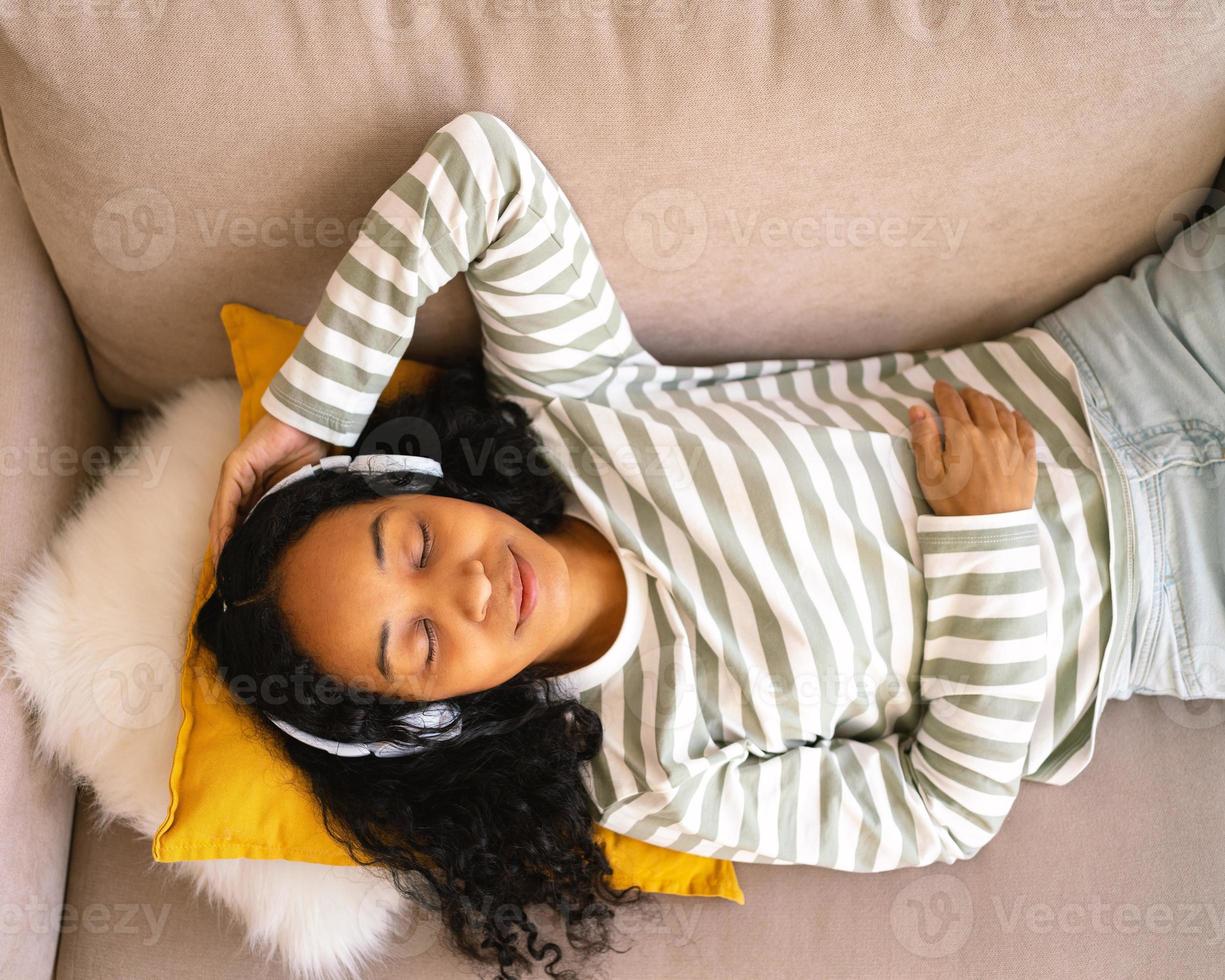 Calm african-american female lying on couch, listening to music in headset. Enjoying audio playing photo