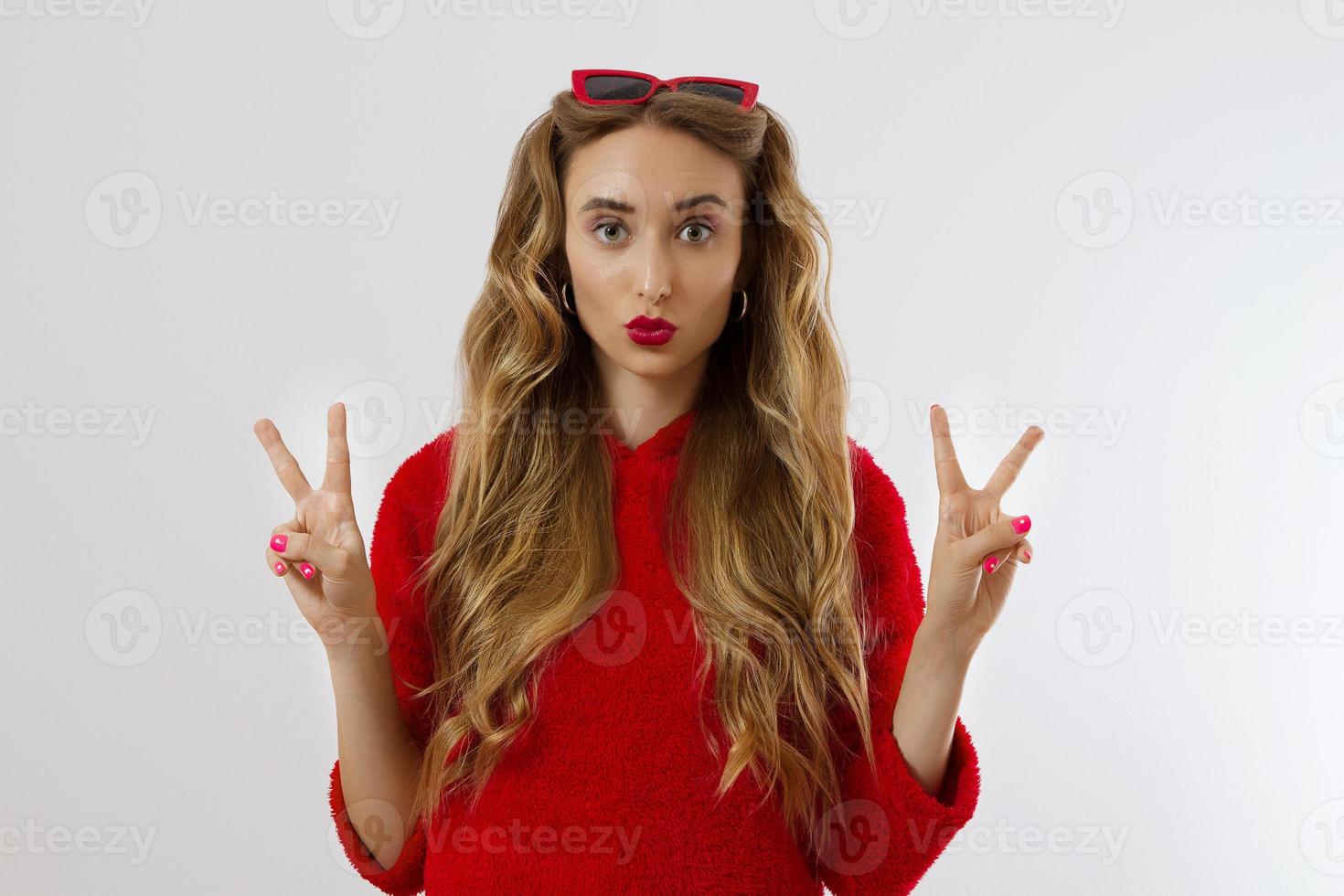 Young woman in red hoodie sweatshirt isolated on white background. Fashion and style concept. Peace sign gesture. Happy beautiful girl in red sunglasses. Make-up and beauty. Glamour model. Kiss face photo