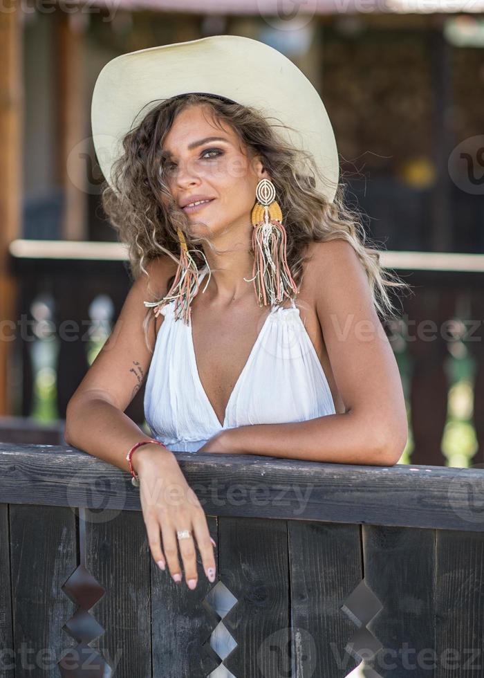 hermosa mujer rubia con cabello rizado y sombrero de vaquero posando. retrato de una chica con vestido blanco en el rancho mirando a la cámara foto