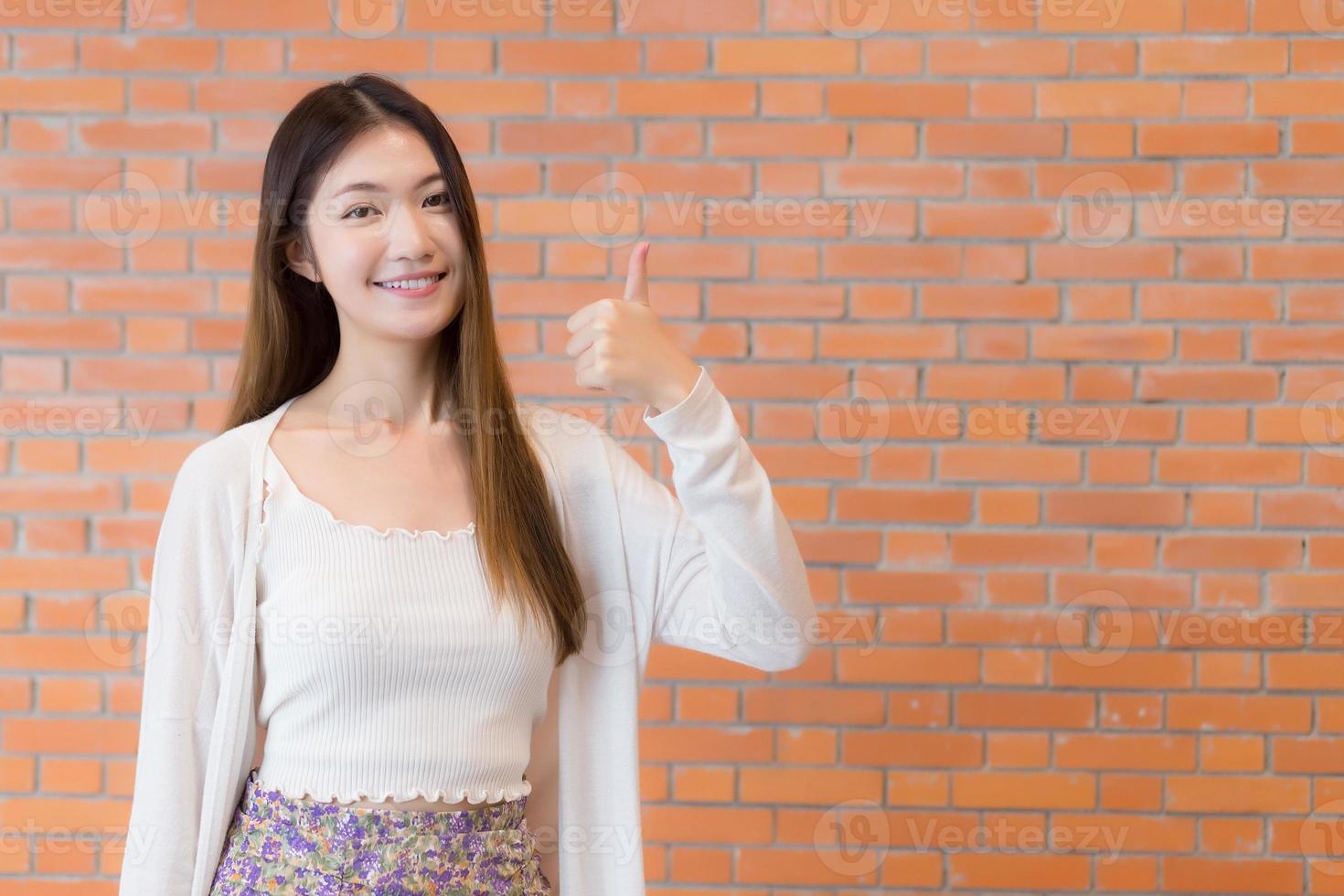 hermosa mujer asiática joven de negocios que tiene el pelo rubio largo con una camisa azul sonríe y muestra el pulgar hacia arriba y presenta algo sobre un fondo de ladrillo marrón. foto