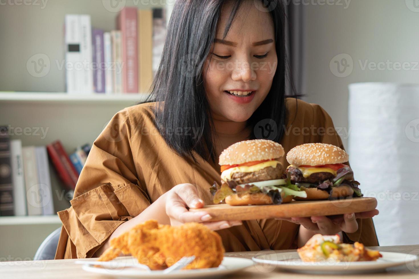 mujer hambrienta con sobrepeso sosteniendo hamburguesa en un plato de madera, pollo frito y pizza en la mesa, durante el trabajo desde casa, aumenta el problema de peso. concepto de cama de trastorno por atracón compulsivo foto