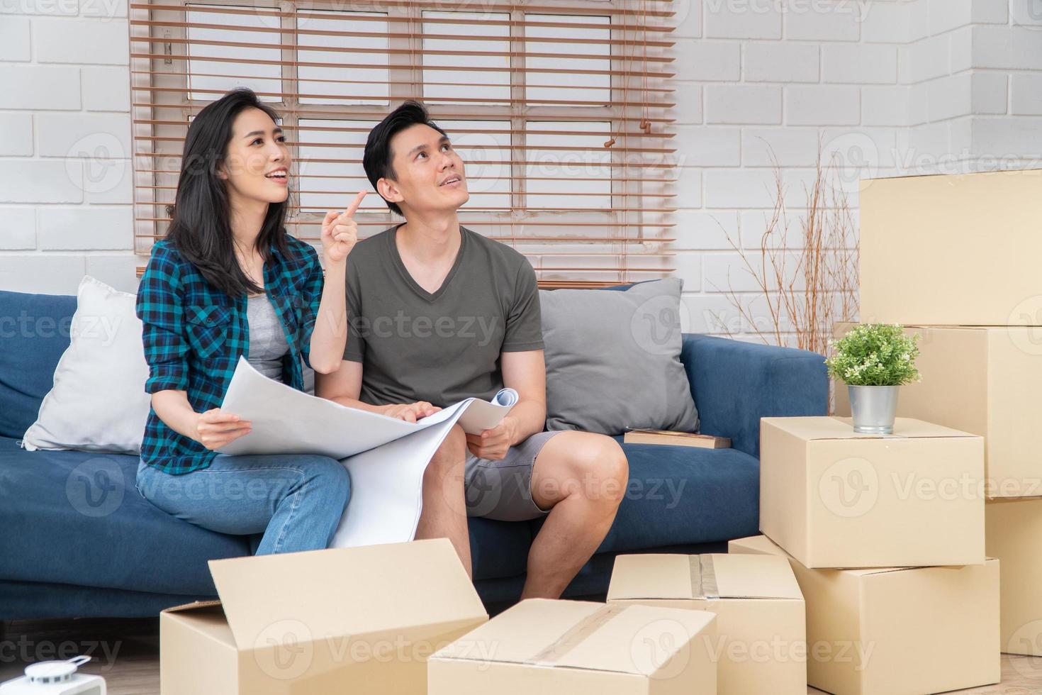 Smiling young Asian happy couple hold blueprint for home decoration ideas at moving day in their new home after buying real estate. Concept of starting a new life for a newly married couple. photo