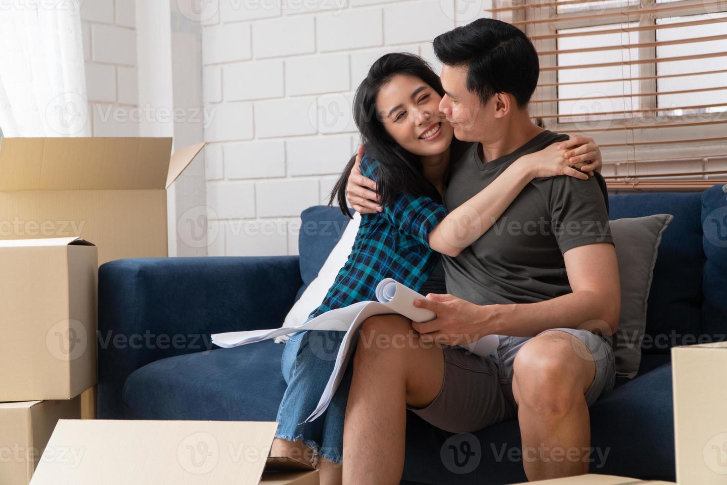 Smiling young Asian happy couple hold blueprint for home decoration ideas at moving day in their new home after buying real estate. Concept of starting a new life for a newly married couple. photo