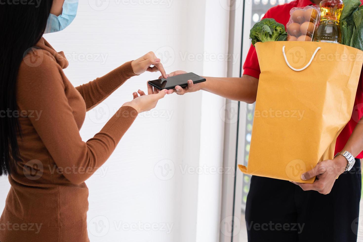 Asian delivery man holding holding a bag of fresh food for giving to customers and holding smartphone for Receive payments at home. Concept of express grocery service and new lifestyle photo