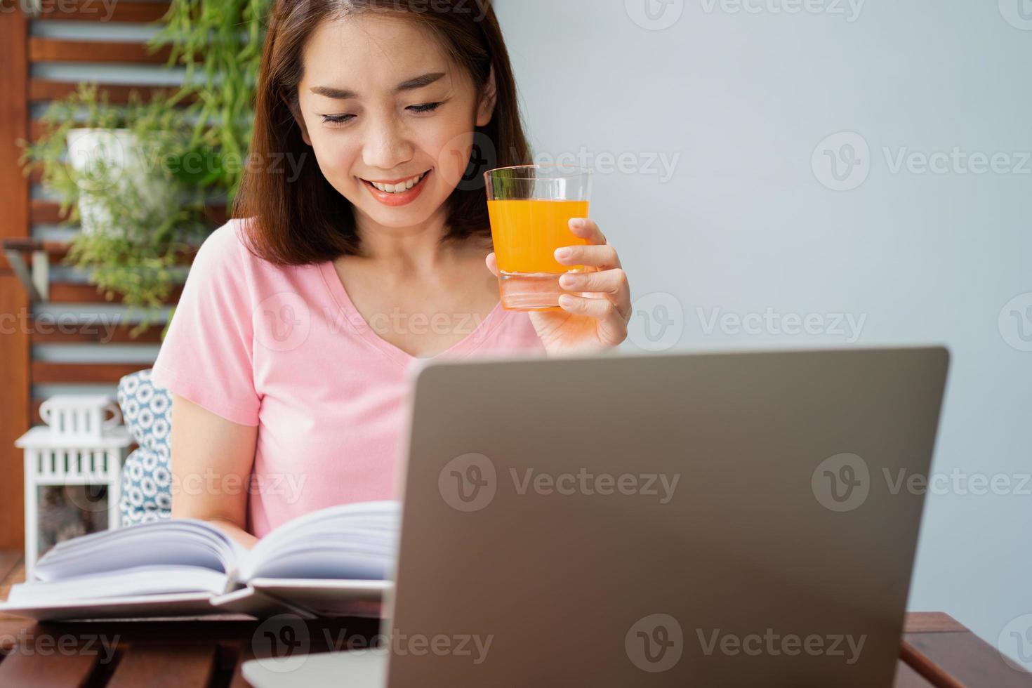 mujer asiática de mediana edad leyendo un libro y sosteniendo un vaso de jugo de naranja en el hogar. concepto de cuidado de la salud y alimentación saludable foto