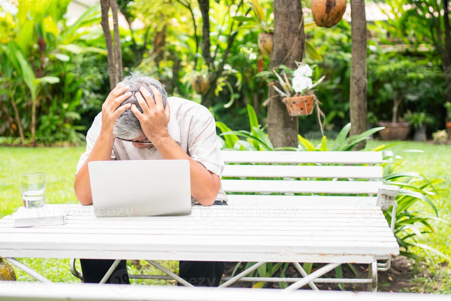 Portrait of an old elderly Asian man holds the head with his hand cause of stress after try to use a computer laptop in the backyard after retired. Concept of Ageism and Hobbies after retirement. photo
