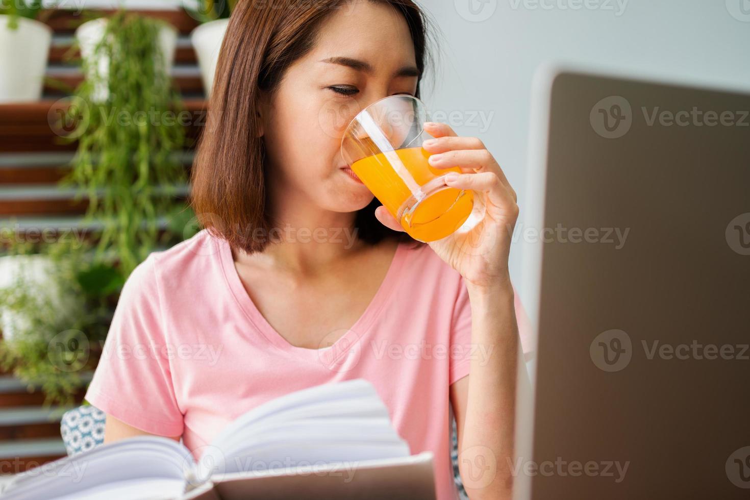 mujer asiática de mediana edad leyendo un libro y sosteniendo un vaso de jugo de naranja en el hogar. concepto de cuidado de la salud y alimentación saludable foto