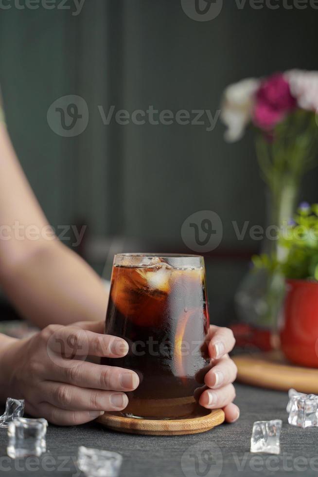 Hands holding a glass of espresso with lemon juice and fresh sliced lemon on wooden table and copy space, Summer Cocktail, Cold brew coffee or black tea. photo