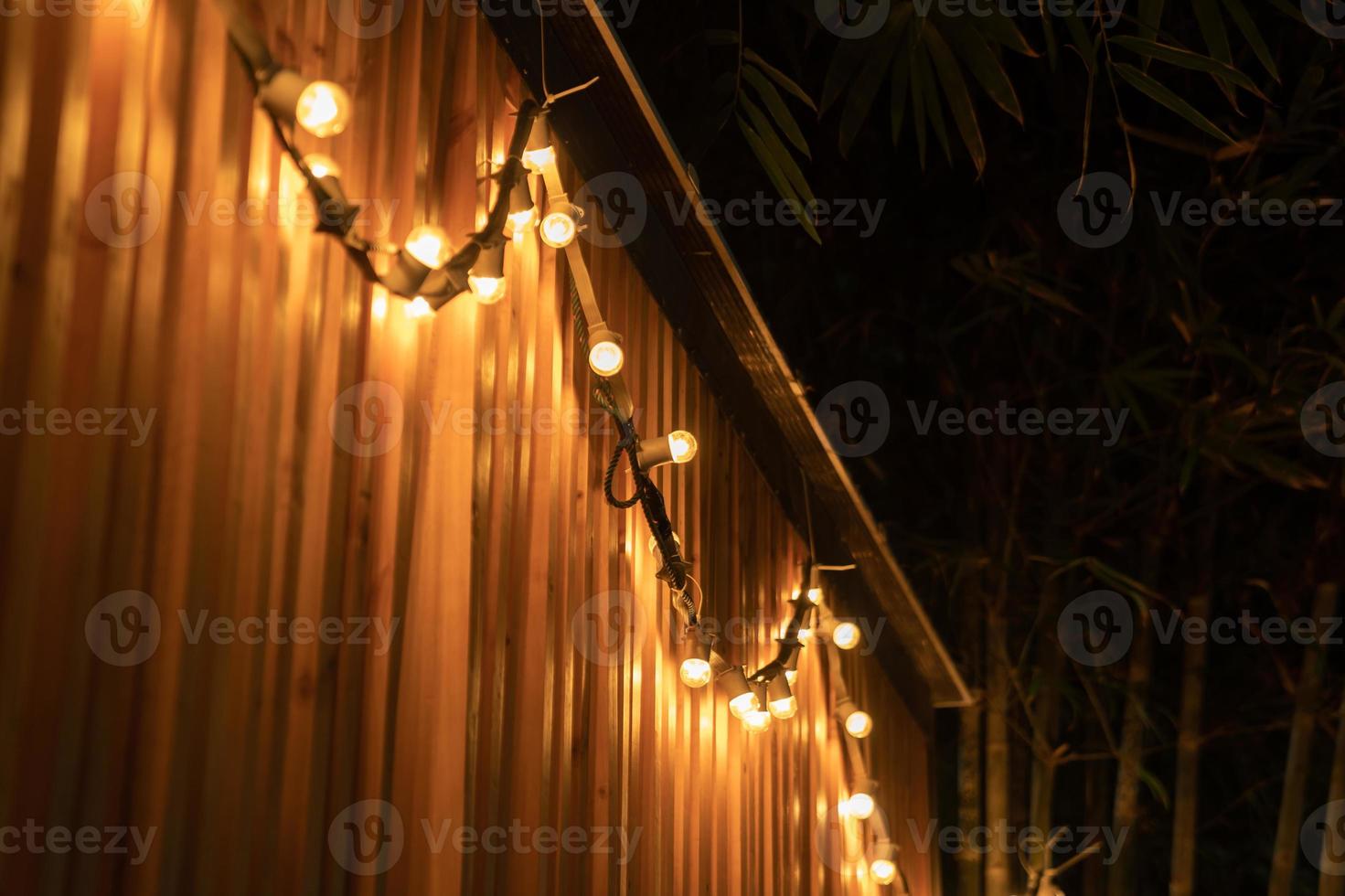 la luz de la lámpara de bola de tungsteno en la línea se cuelga en la partición de listones de madera en la noche al aire libre con fondo de árbol de bambú. foto