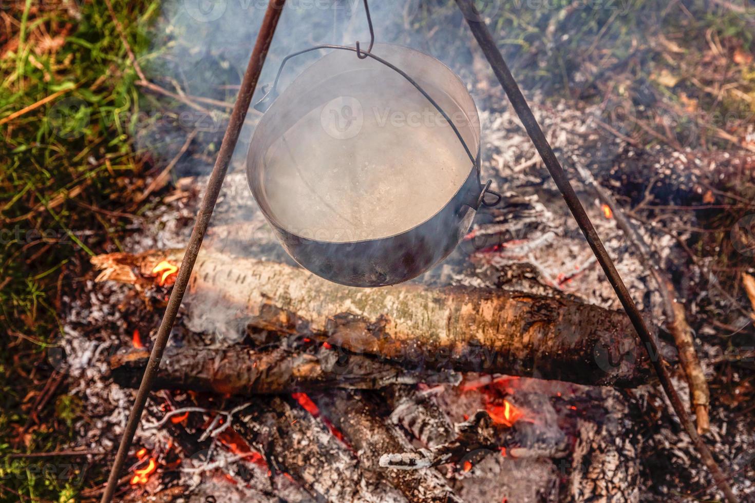 Cast iron pot cooks over open fire in a campsite in forest in sunny summer day. Camping tourism relax trip active lifestyle adventure travel picnic concept. photo