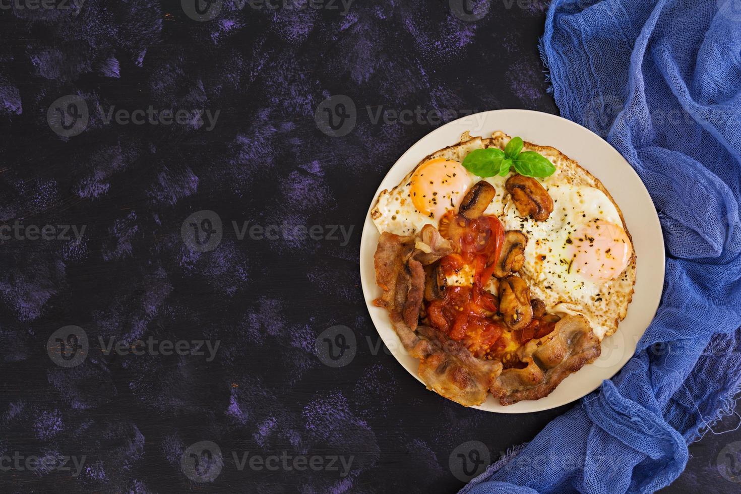 Fried eggs with bacon, tomatoes and mushrooms on dark background. Top view photo