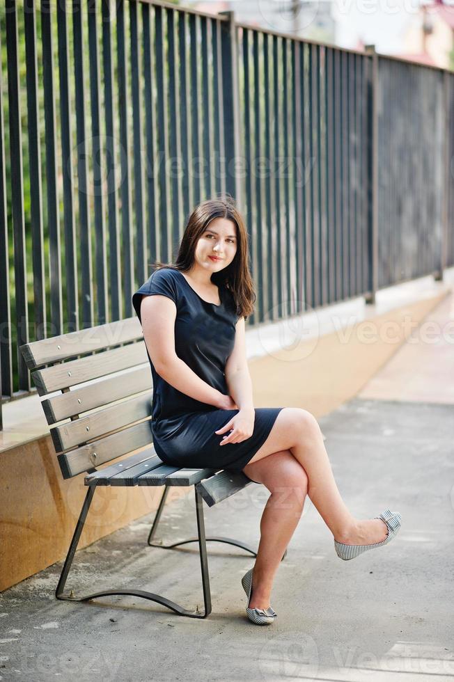 chica morena con vestido negro sentada en un banco y posando en la calle de la ciudad. foto