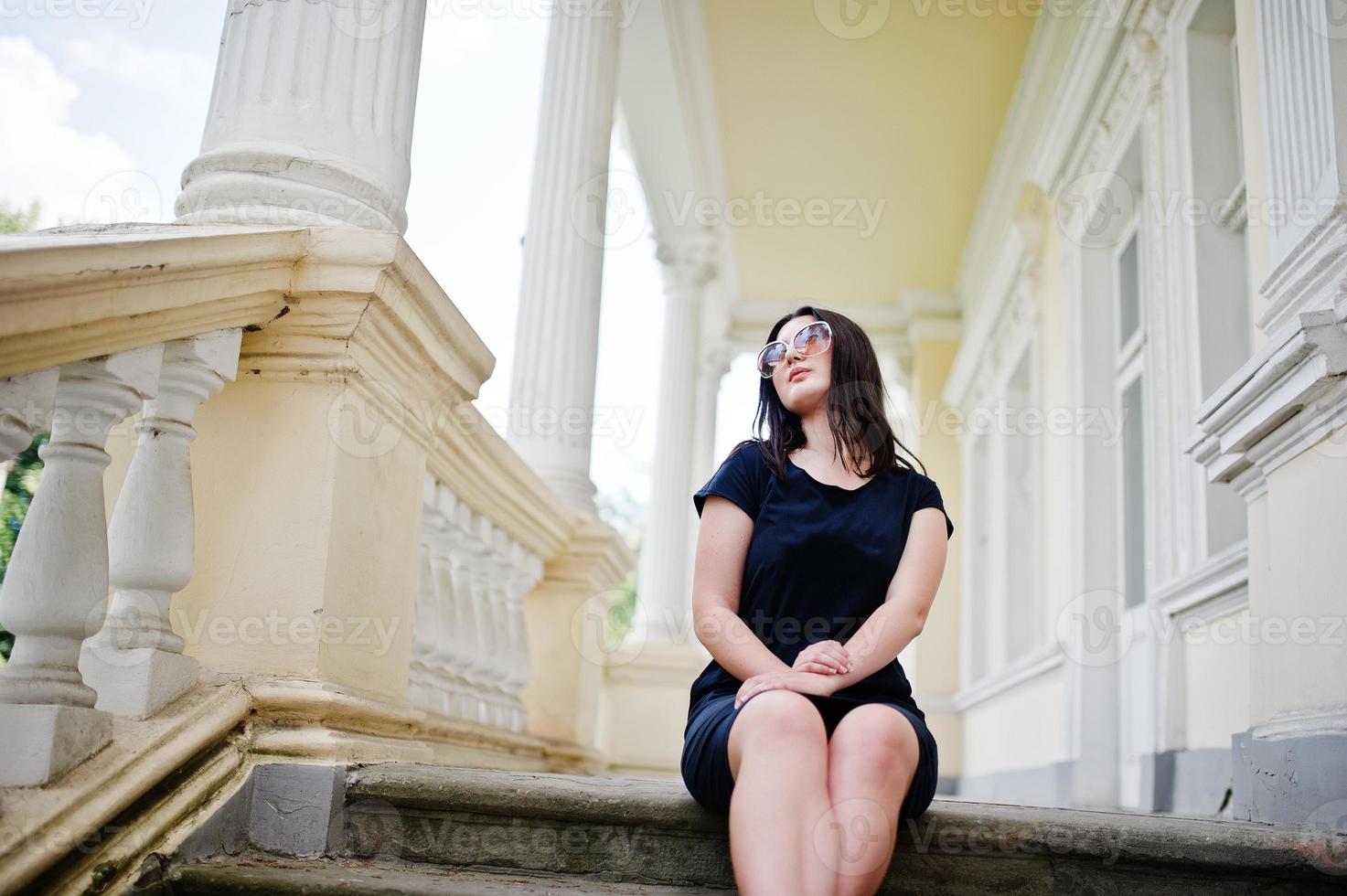 Brunette girl at black dress, sunglasses sitting on stairs of old vintage house, posing at street of city. photo